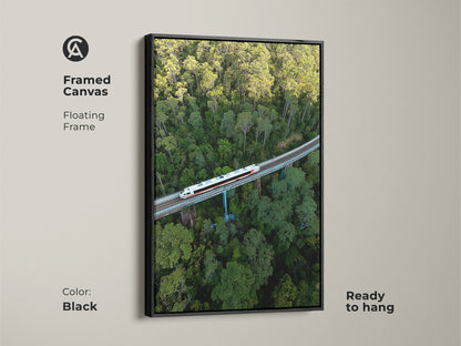 Framed canvas art showing an aerial view of a train. The artwork has a floating frame in black. The green forest canvas is ready to hang. This modern transportation print enhances living room art. The railway bridge artwork captures a green toned wall art.