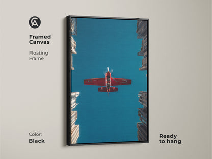 Framed canvas print of a red airplane flying over city skyscrapers. The floating frame is black. This aviation artwork offers a contemporary feel. Ready to hang. A perfect gift for pilots or aviation art lovers. The sky is deep blue.