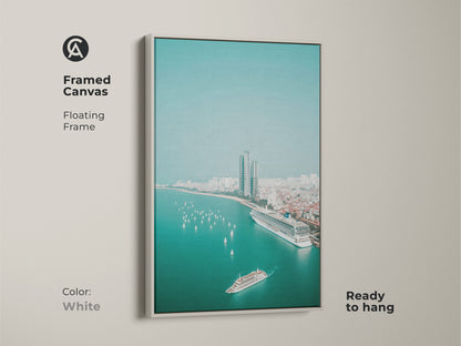 Framed canvas with a white floating frame displays teal ocean cityscape art. An aerial view includes a cruise ship sailboats and city buildings. This contemporary artwork is ready to hang as office cityscape art.