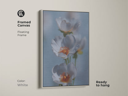 Framed canvas print of delicate white flower artwork. This soft focus botanical art is in a white floating frame. The light blue floral canvas complements minimalist decor. This gentle flower art print adds a calming touch. It makes a wonderful housewarming gift for new homeowners.