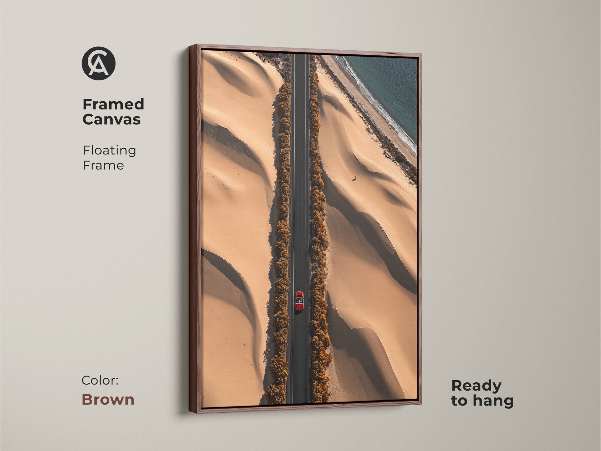 This desert road art print is framed in brown. This aerial canvas features a red car on a sandy beige road. The contemporary travel landscape offers an ocean view. Ready to hang this artwork brings a modern aesthetic to your home. Perfect for travel enthusiasts. This is a unique road trip gift idea.