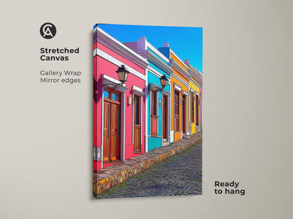A gallery-wrapped stretched canvas shows a cartoonish cityscape art print. Colorful houses in hot pink sky blue and teal line a cobblestone street. It's ready to hang as eclectic home decor for travel lovers. It adds vibrant character to any living space. This European street scene is a unique and eye-catching wall art.