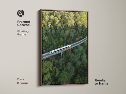 Framed canvas art displays an aerial train view. This modern artwork features a floating frame in brown. A green forest canvas is ready to hang. The contemporary train art is ideal for bedroom landscape art. This nature landscape print is a travel enthusiast gift.