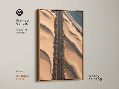 A framed desert road art print in antique gold showcases a red car on a sandy beige road. This contemporary travel landscape art captures dunes meeting the ocean. Ready to hang this artwork adds a touch of modern desert art to your space. It is a unique home art piece for your living room.