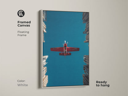 Framed canvas aviation artwork with a white floating frame. A crimson red aeroplane flies through a deep blue sky above urban skyscrapers. Ready to hang. This aviation photography print is a great office decor or a pilot gift idea.