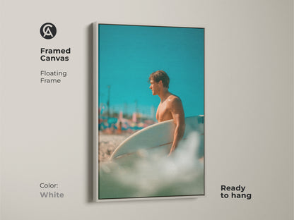 Framed canvas artwork of a surfer on a beach. The floating frame is white. The art shows a man holding a surfboard. This ready-to-hang framed canvas will add a coastal vibe to any space.