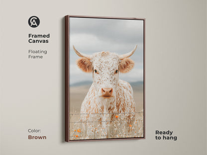 Framed canvas artwork of a white and brown speckled longhorn cow in a brown floating frame. The cow is standing behind a barbed wire fence. Small orange wildflowers grow at the base. This framed art is ready to hang.