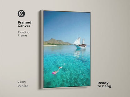 A white framed canvas features a sailboat on clear turquoise water. This coastal artwork shows a snorkeler enjoying the ocean. The floating frame complements any room. Ready to hang and enjoy.
