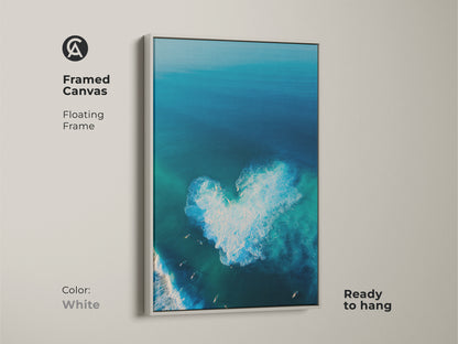 White framed canvas art showing a serene aerial ocean scene. A heart-shaped wave is visible. Surfers are enjoying the surf. This floating frame canvas is ready to hang. The white frame offers a clean modern look. The artwork brightens any space. 