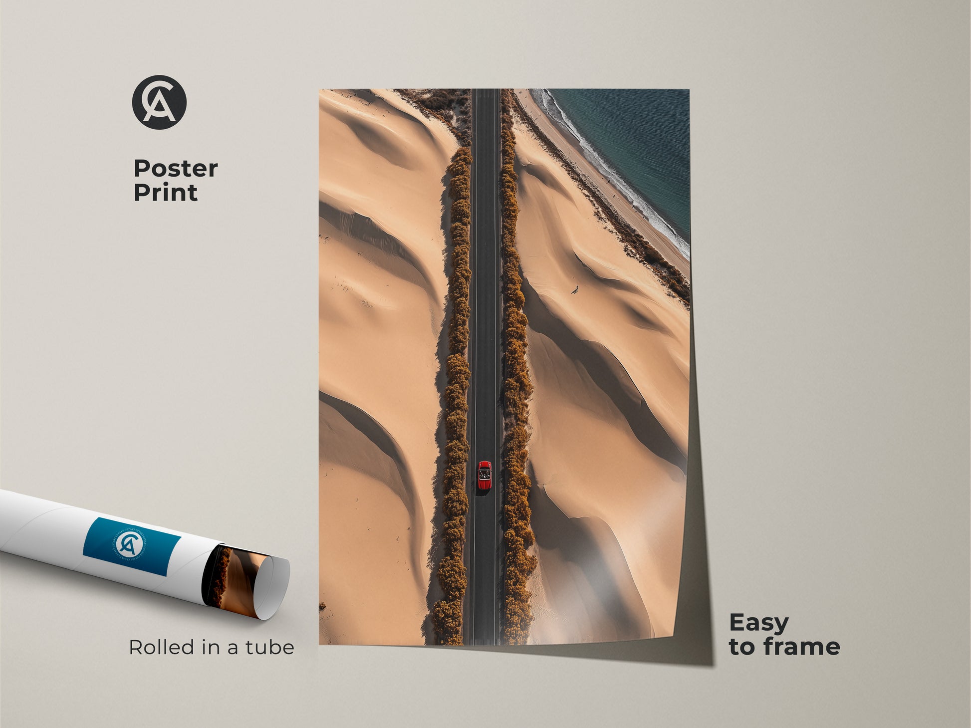 A contemporary aerial desert road art poster print shows a red car on a sandy beige road. This travel adventure art piece is easy to frame. It captures a stunning landscape with dunes and an ocean view. Rolled in a tube for safe shipping. This modern desert art is perfect for hallway wall art.