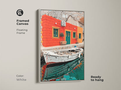 The white framed canvas displays a vibrant Mediterranean scene. An orange teal house is showcased with coastal boat artwork. This realism landscape serves as a thoughtful housewarming gift.