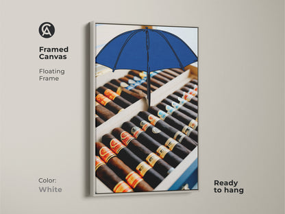 White framed canvas print with a cigar box and blue umbrella. The pop art piece offers a clean modern look. Enhance your space with this office decor canvas art. Ready to hang for instant style. The tobacco art is perfect for contemporary and eclectic interiors.