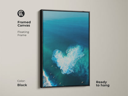 Black framed canvas art brings a modern touch to any room. An aerial ocean scene with a heart-shaped wave. Surfers are below. This floating frame canvas is ready to hang. The black frame enhances the vibrant colors of the ocean. The artwork is eye-catching.