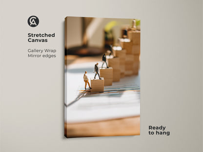 This stretched canvas features miniature figures climbing wooden blocks gallery wrapped with mirror edges. This office wall art symbolizes progress. The conceptual business print is a motivational piece for any ambitious professional. It's ready to hang.