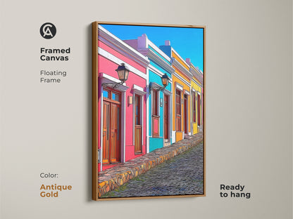 Framed canvas art shows a cartoonish cityscape with colorful houses and a cobblestone street. An antique gold floating frame highlights this unique wall decor. The hot pink sky blue and teal buildings give an eclectic feel. Perfect for a living room this ready to hang European street scene is a vibrant gift for travel enthusiasts.