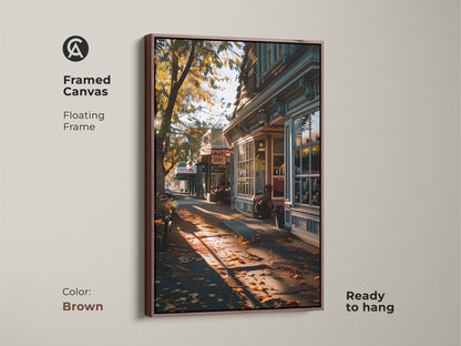 Framed canvas print capturing an autumn street view. Brown floating frame complements the traditional storefront art. Golden hour light adds warmth and charm. Ready to hang for instant display. Ideal for fall season wall decor. This is cozy and inviting art.