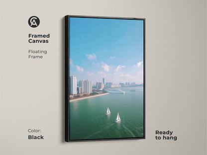 Framed canvas art featuring an aerial cityscape with sailboats. This ocean view artwork has a black floating frame. The modern travel art is ready to hang showcasing a city skyline in teal blue. This coastal decor canvas brings a contemporary style to any room. The artwork is perfect for adding nautical home decor.