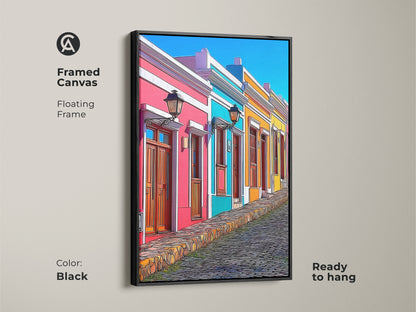 Framed canvas art featuring a cartoonish cityscape with colorful houses and a cobblestone street. A black floating frame enhances this vibrant wall art. The hot pink sky blue and teal buildings create an eclectic feel perfect for a living room. Ready to hang this European street scene makes a unique travel lover gift.