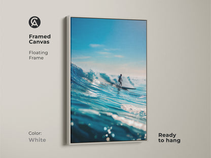 A framed canvas artwork showcases a surfer riding a magnificent blue wave. The white floating frame offers a clean and modern look. Ready to hang this print brings the energy of the ocean indoors. An excellent addition to any surf art collection.