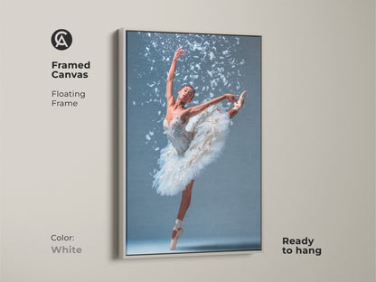 Framed canvas art featuring a ballerina. She is wearing a white tutu. She is en pointe with one leg extended. White feathers accent the artwork. The floating frame is white. This beautiful ballet art is ready to hang.
