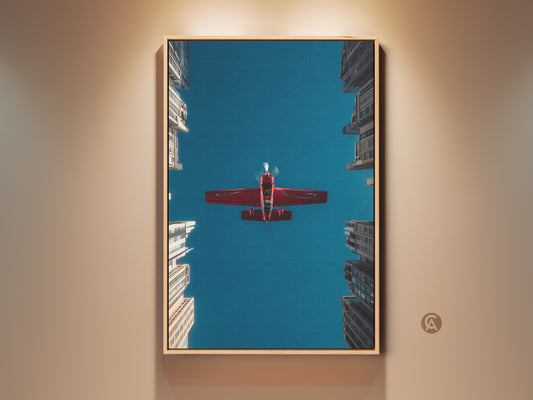 Aviation art print featuring a crimson red aeroplane soaring through a deep blue sky. Urban skyscrapers frame the shot. This urban photography wall decor is perfect as office artwork or a pilot gift. The contemporary aviation artwork is framed in oak.