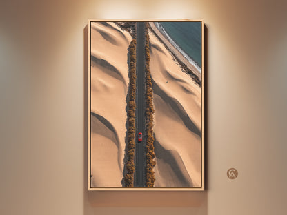 This aerial desert road art print features a red car driving on a sandy beige road. The contemporary travel artwork captures a winding road between dunes and an ocean view. A perfect hallway decor piece. This canvas print brings a modern landscape photography aesthetic to your home or office. It is a unique gift for travel enthusiasts.