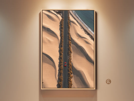 This aerial desert road art print features a red car driving on a sandy beige road. The contemporary travel artwork captures a winding road between dunes and an ocean view. A perfect hallway decor piece. This canvas print brings a modern landscape photography aesthetic to your home or office. It is a unique gift for travel enthusiasts.