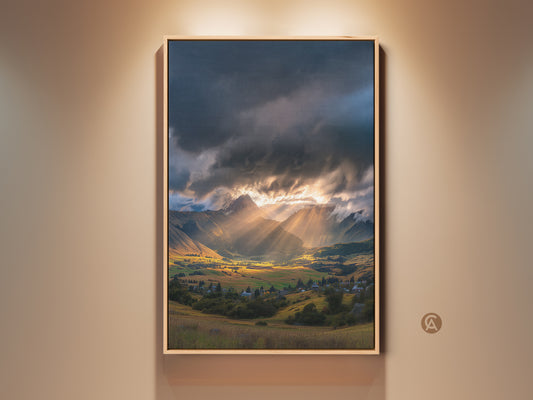 Framed mountain valley canvas art featuring golden hour sun rays. Dramatic sun rays pierce through cloudy skies over a picturesque village. This contemporary nature decor is perfect for adding a serene landscape print to any room.
