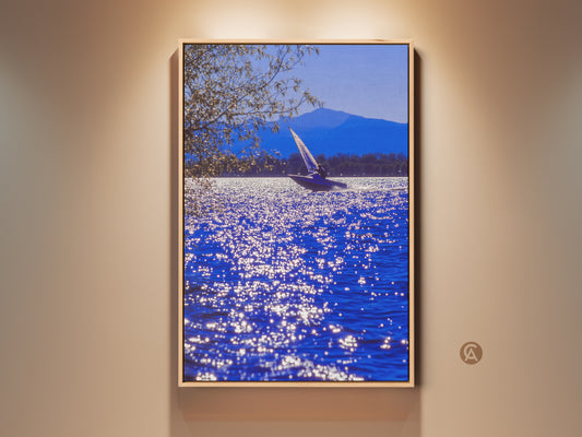 Realistic sailboat art print displayed in a residential setting. The deep blue water sparkles. A mountain range is visible in the distance. It is a contemporary coastal art piece for home or office spaces.