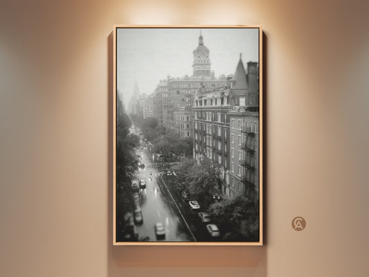 Black and white photograph of a city street. The artwork shows a view from above. Cars are driving on a wet road. Buildings line both sides of the street. This framed canvas art is ready to hang.