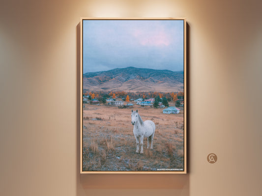 Framed fine art showing a white horse in a field. A village sits at the base of distant mountains under a cloudy sky. The artwork is printed on high-quality canvas and framed in a natural wood.