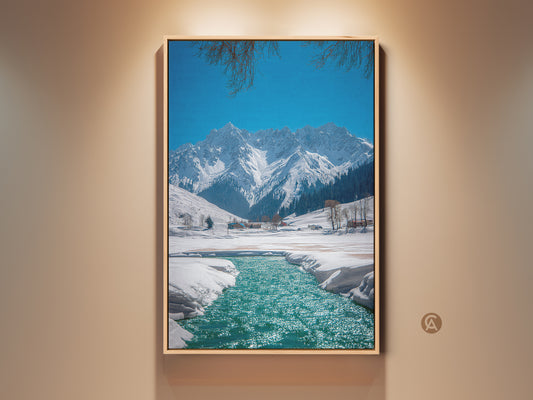 Rustic winter landscape artwork displays snow-capped mountains and a teal blue river. A scenic canvas print of nature with a clear blue sky and branches at the top of the frame. Perfect nature wall decor for outdoor enthusiasts a peaceful mountain view.