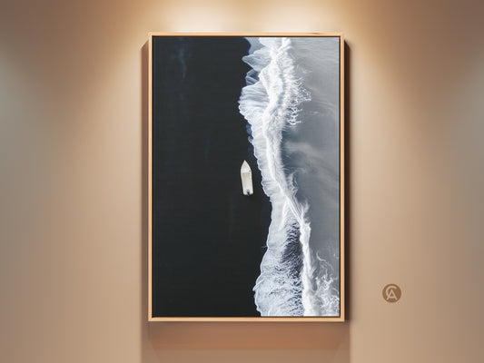 Black and white ocean canvas art in a wood frame hangs on a beige wall. The minimalist boat aerial view is a modern coastal beach wall decor. Perfect bedroom art print.