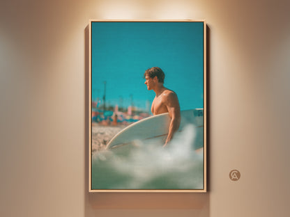 Framed artwork featuring a surfer. The surfer is holding a surfboard on a sunny beach. The artwork is brightly lit. The frame around the art is oak color. The perfect piece for a beach lover.